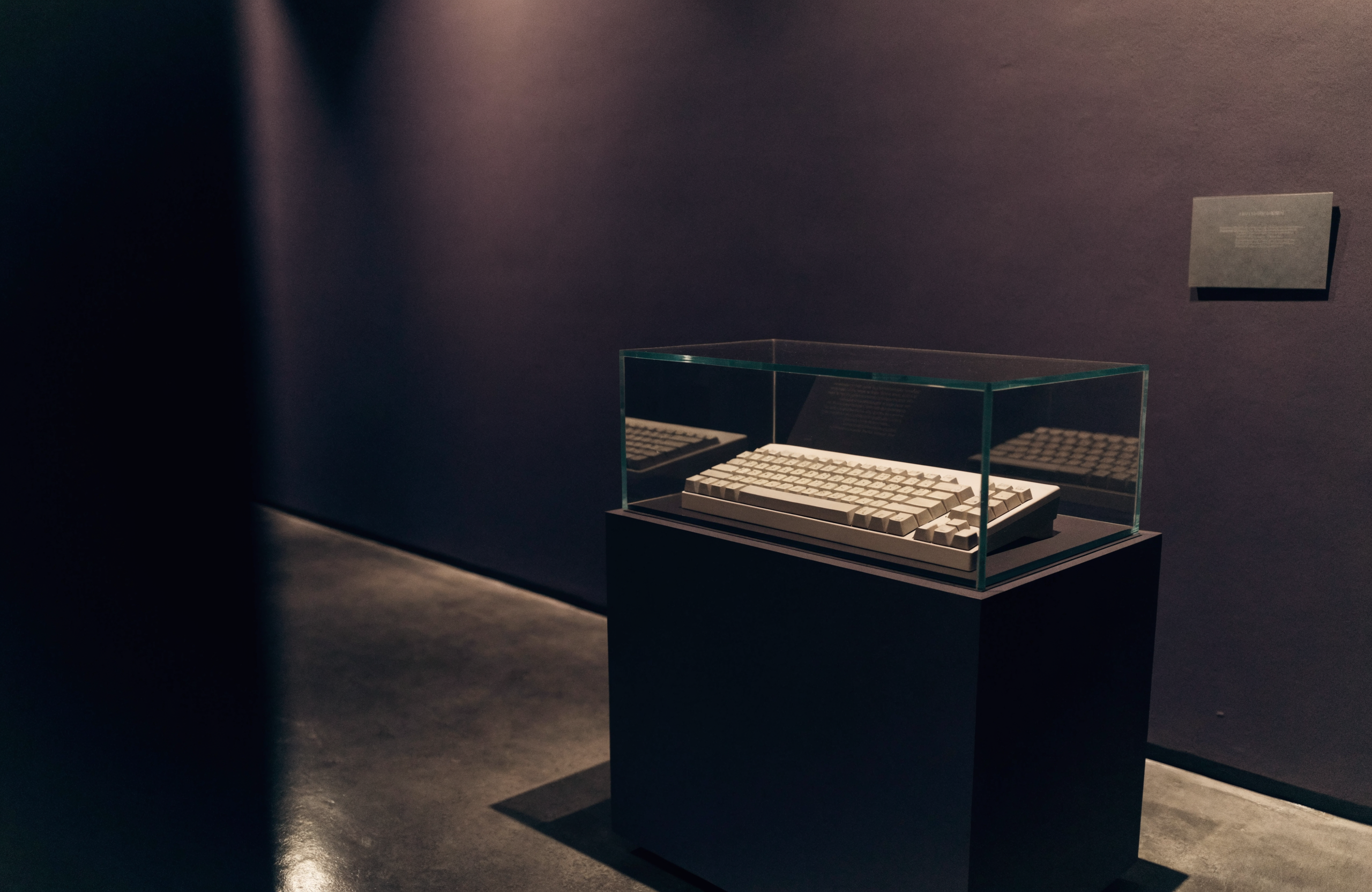 A museum vitrine in a dim gallery: a vintage beige keyboard in a glass case on a charcoal plinth, lit by a single warm spotlight.