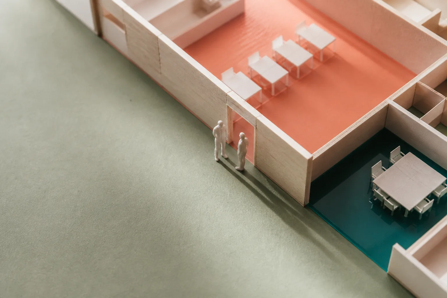 Close photograph of the architectural model: two tiny white figures stand in a doorway cut through a basswood partition between a coral zone with a row of desks and a teal zone with a meeting table